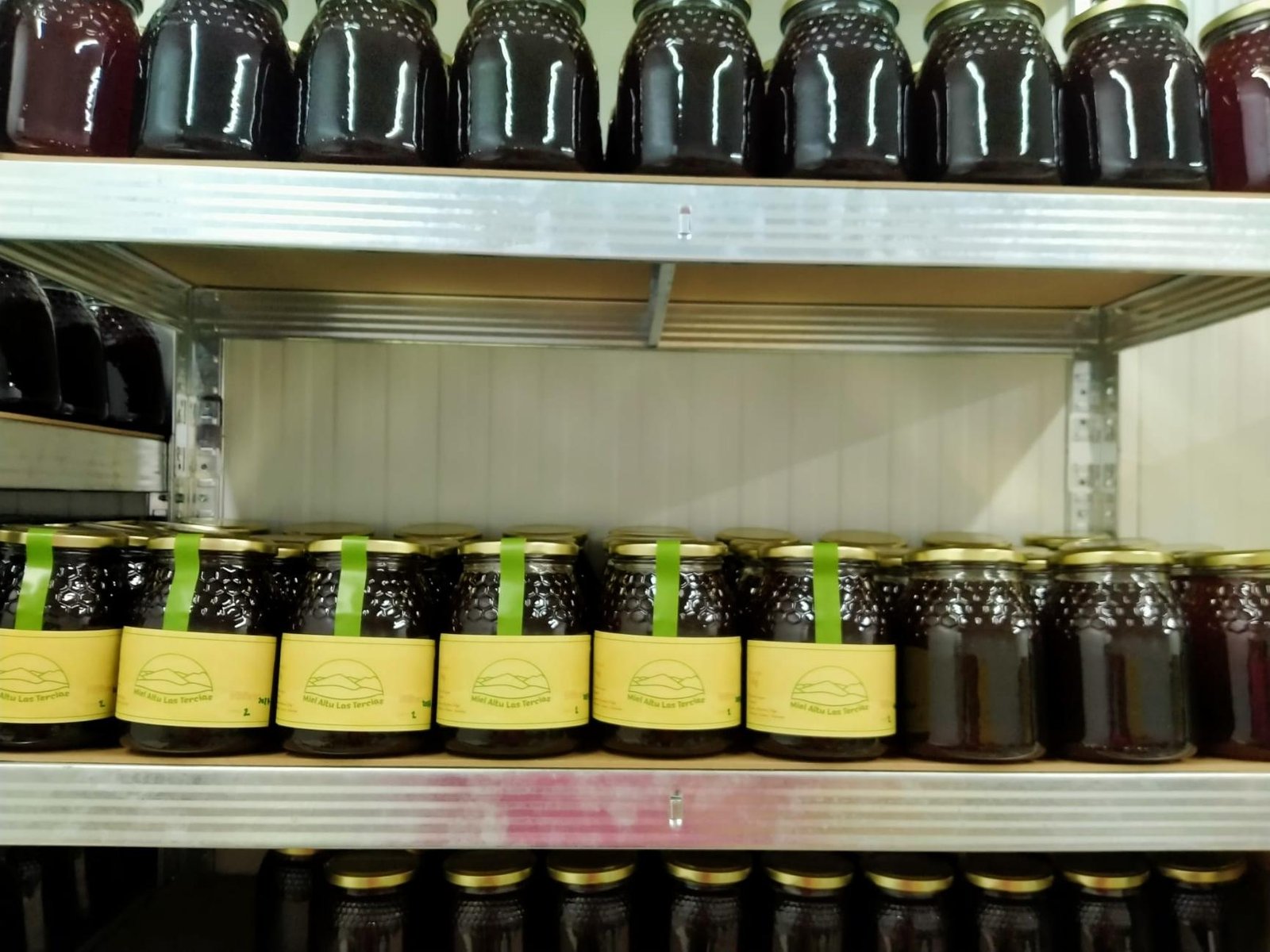 Estanterías con tarros de miel oscura. Varios llevan la etiqueta amarilla y la cinta verde.